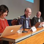 Giacomini, Hartman, and Gupton sit in front of microphones on a conference stage