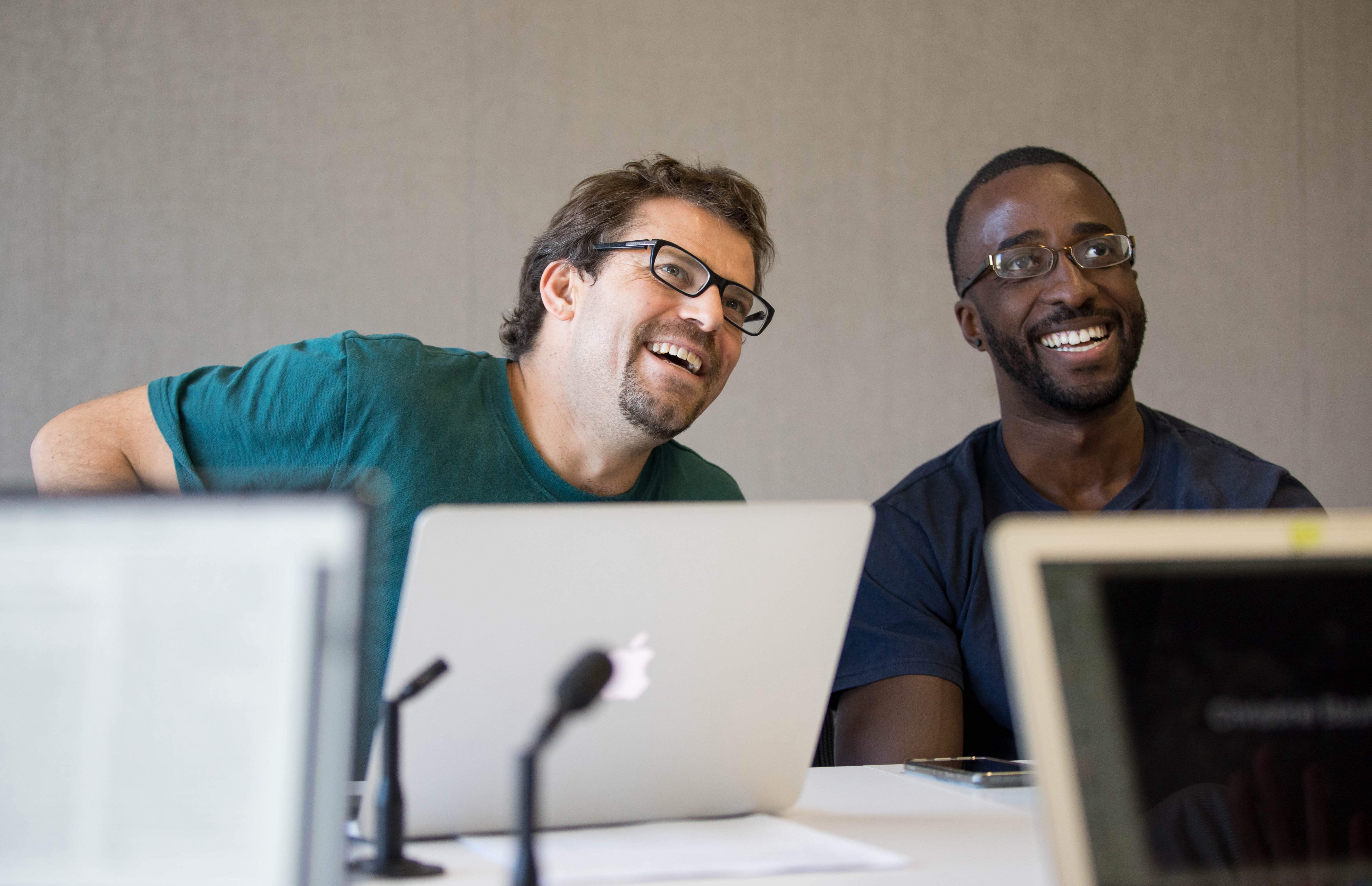 two men with laptops smile in a classroom