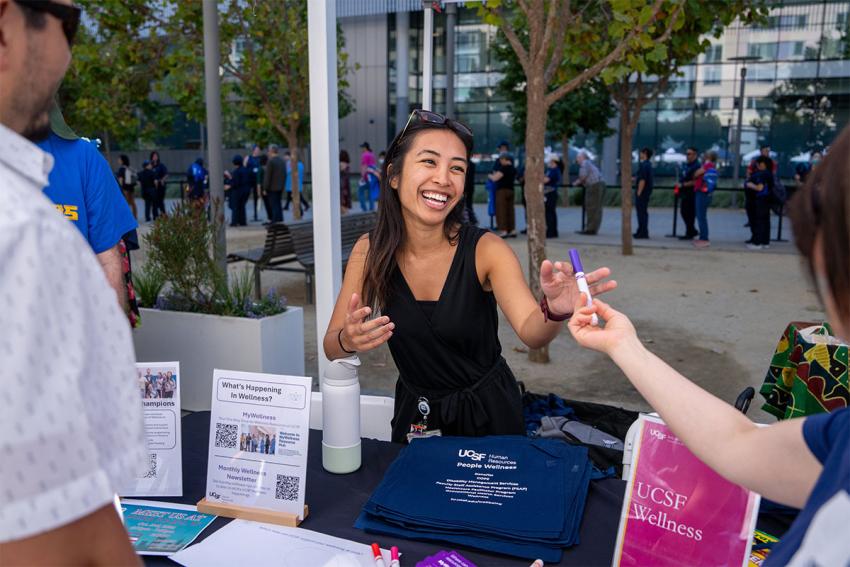 a smiling woman interacts with people at a wellness booth 