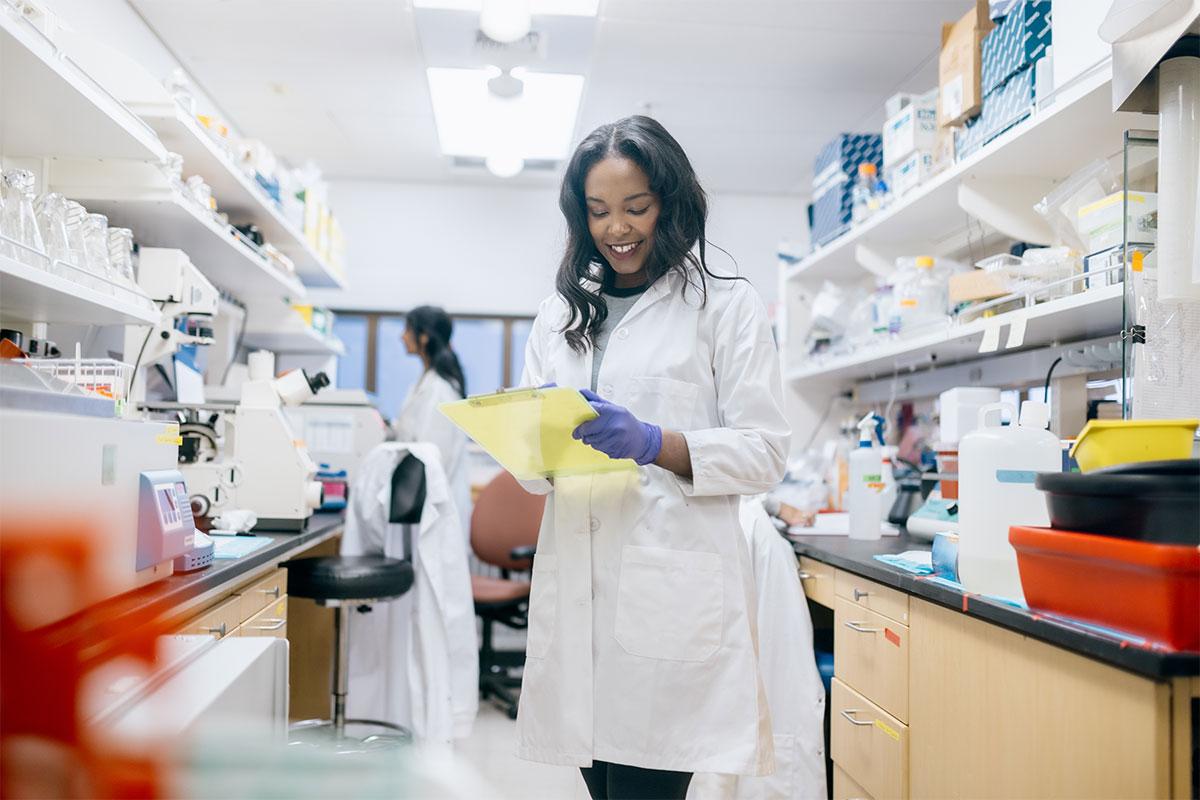 researcher in white lab coat and holding clipboard stands in research lab